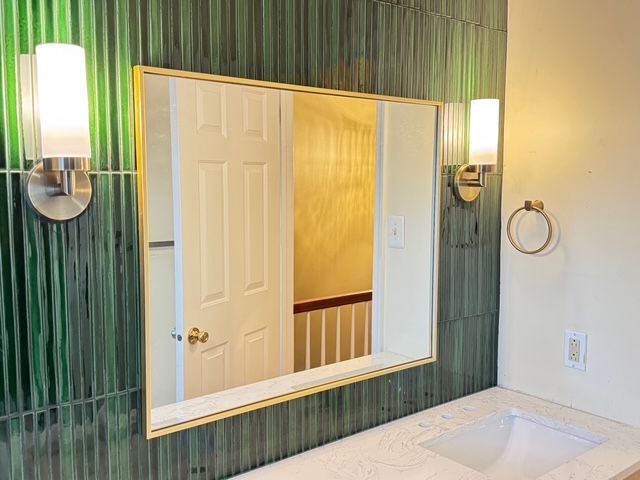 Bathroom vanity with green tile backsplash, gold-framed mirror, sconce lights, and towel ring.