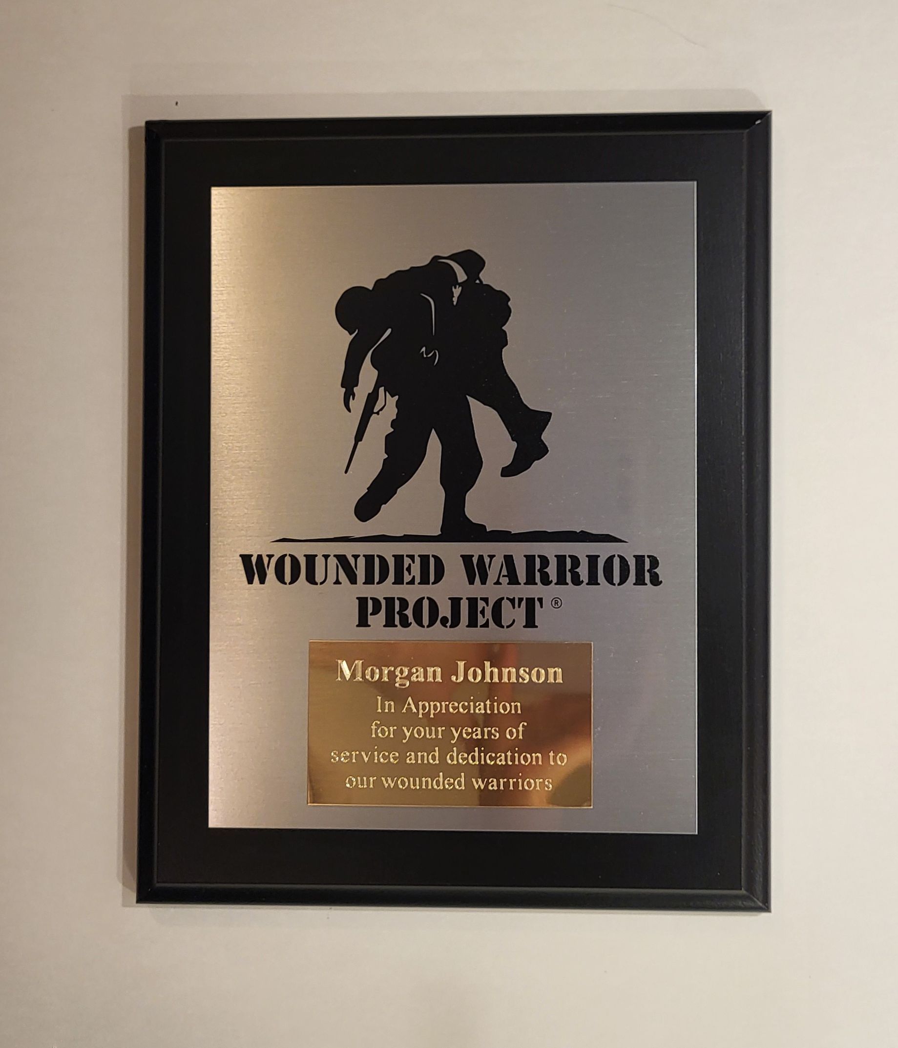 A Wounded Warrior Project plaque featuring a black silhouette of a soldier carrying another, with a gold nameplate below.