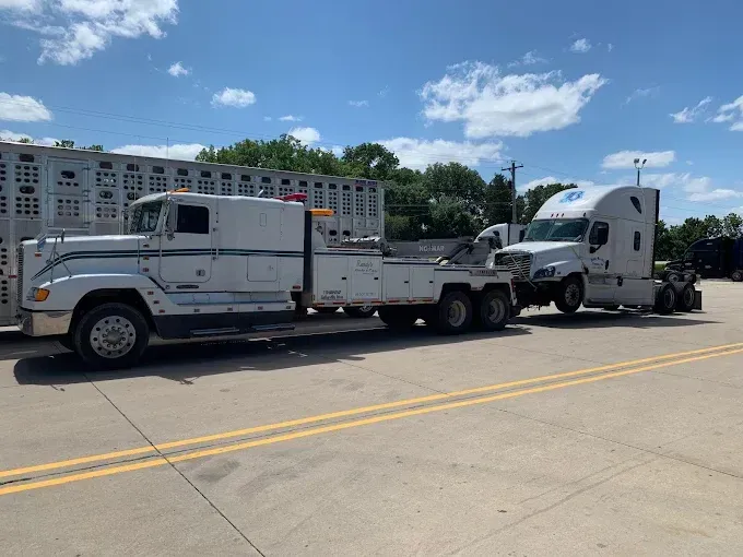 White tow truck towing a white semi-truck on a paved area, under a blue sky.