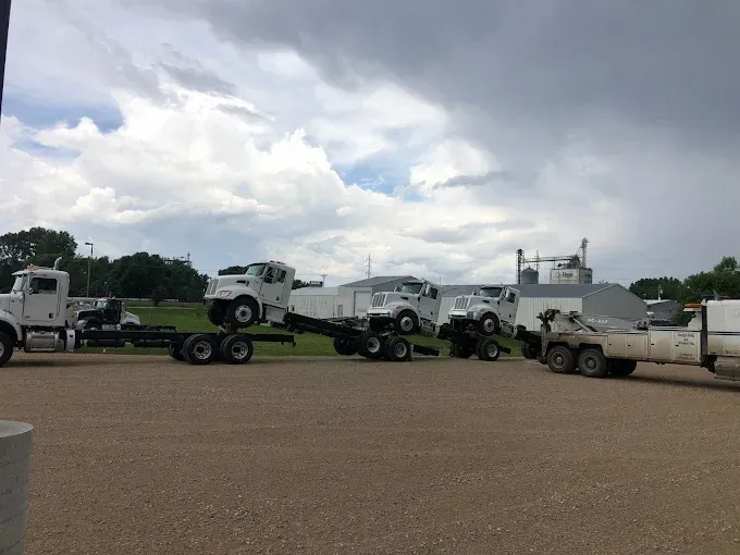 White semi-truck cabs on chassis parked on a gravel lot under a cloudy sky.
