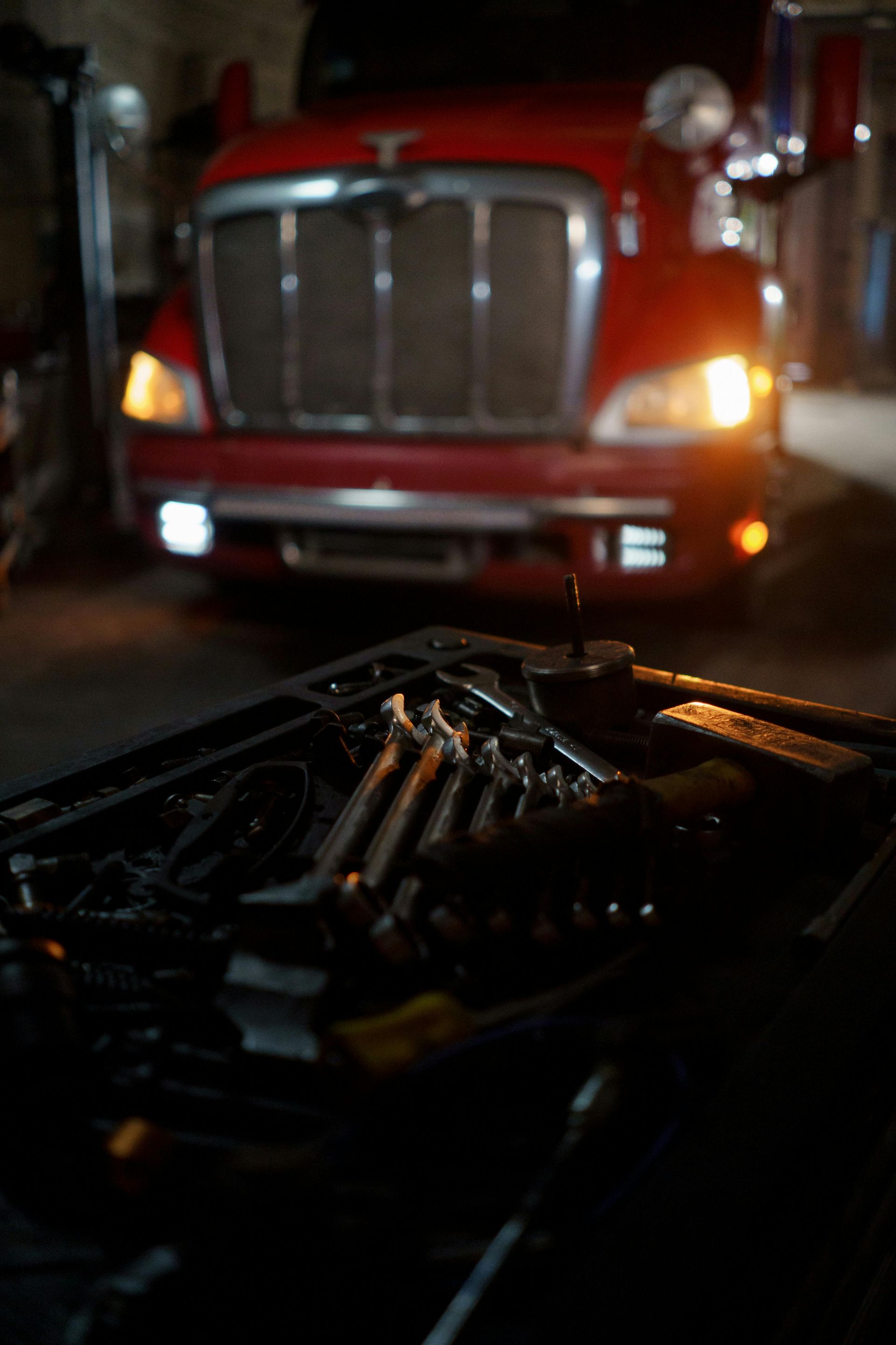 Red semi-truck in a dark garage; tools in foreground. Headlights are illuminated.