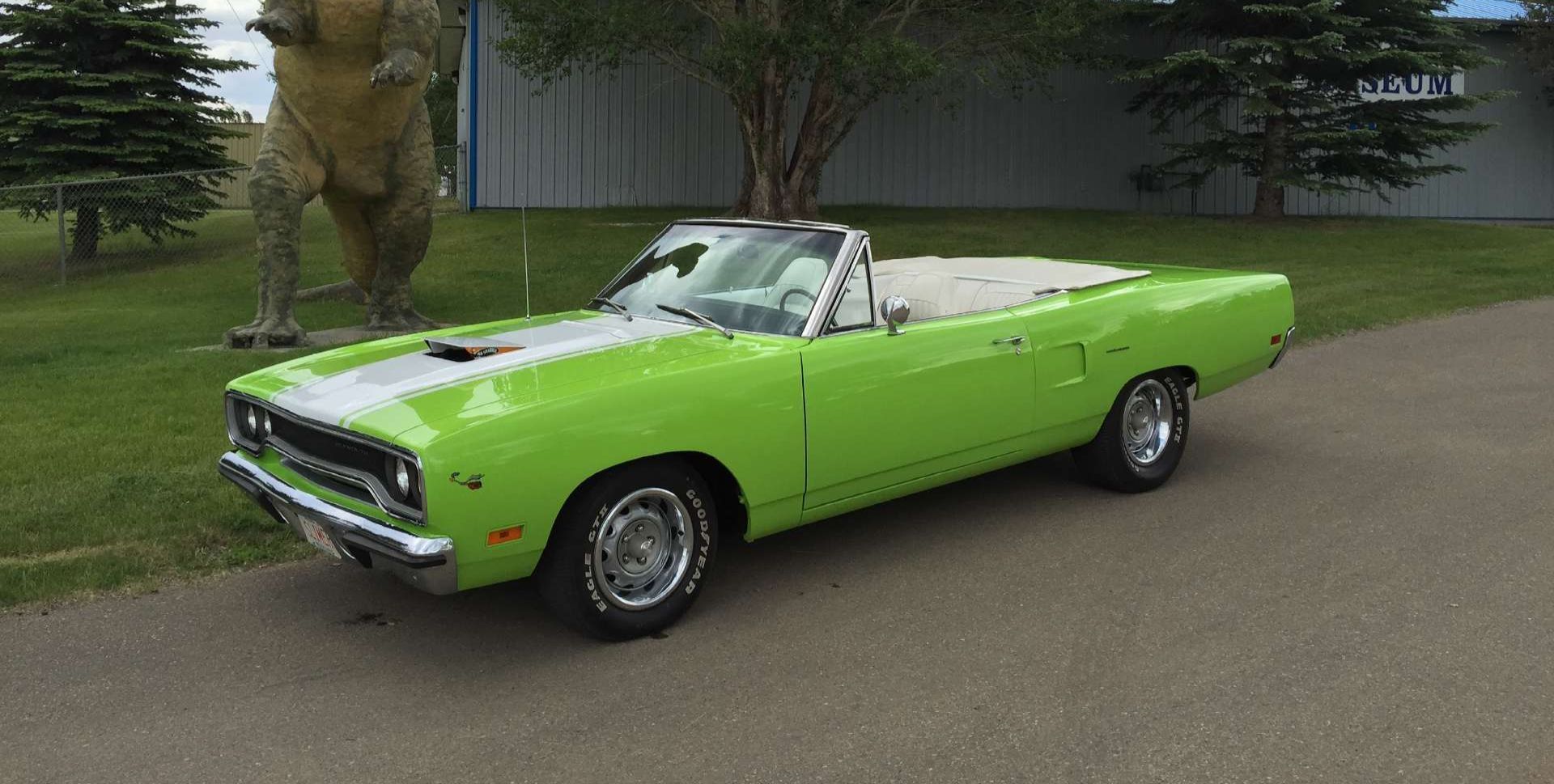 A green convertible dodge plymouth roadrunner is parked in front of a dinosaur statue.
