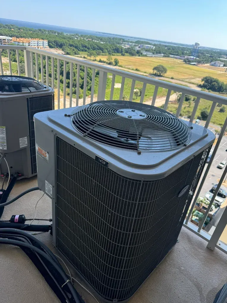 An air conditioning unit on a balcony with a scenic view of buildings and greenery under a blue sky.