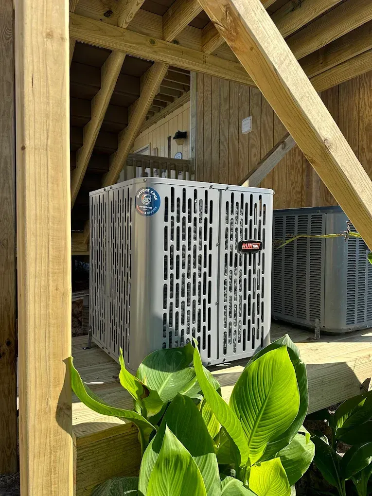 Two air conditioning units under wooden deck, with green plants in foreground.