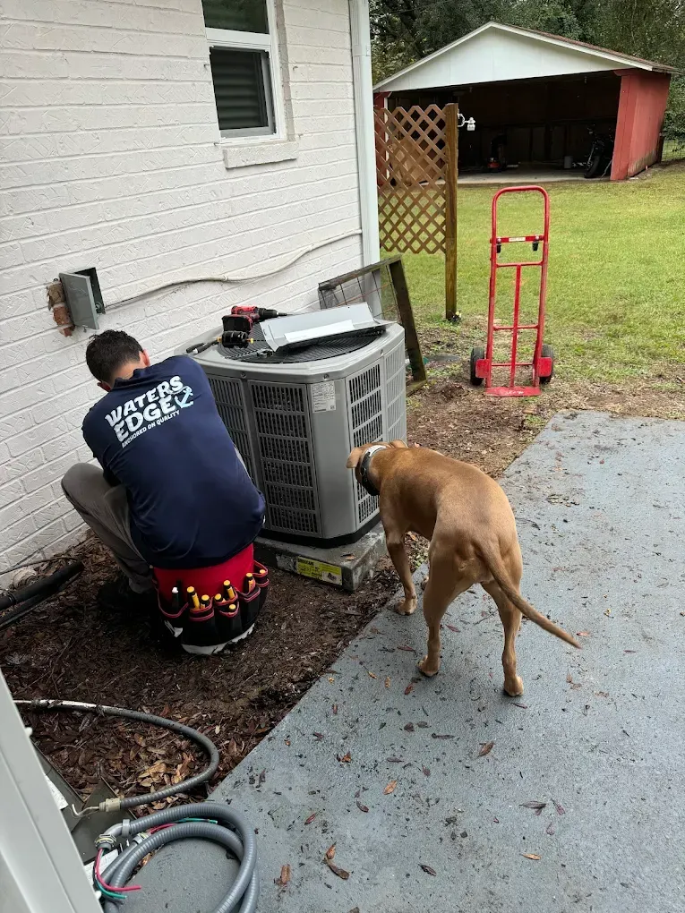 A technician repairs an AC unit outside; a dog sniffs nearby. A hand truck and shed are in the background.