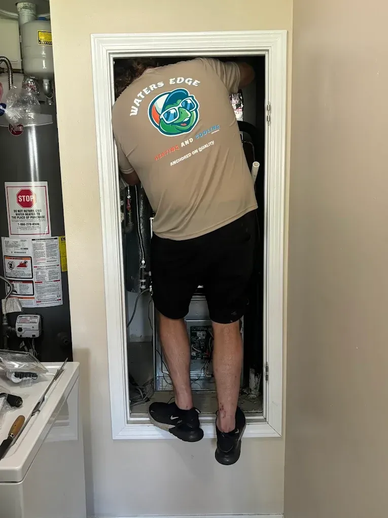 A person in a tan shirt and black shorts is working on equipment in a wall.