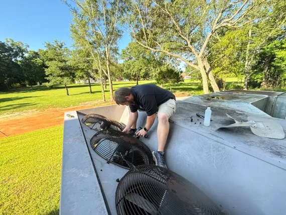 Man working on fans on a concrete structure, outdoors with trees and grass.