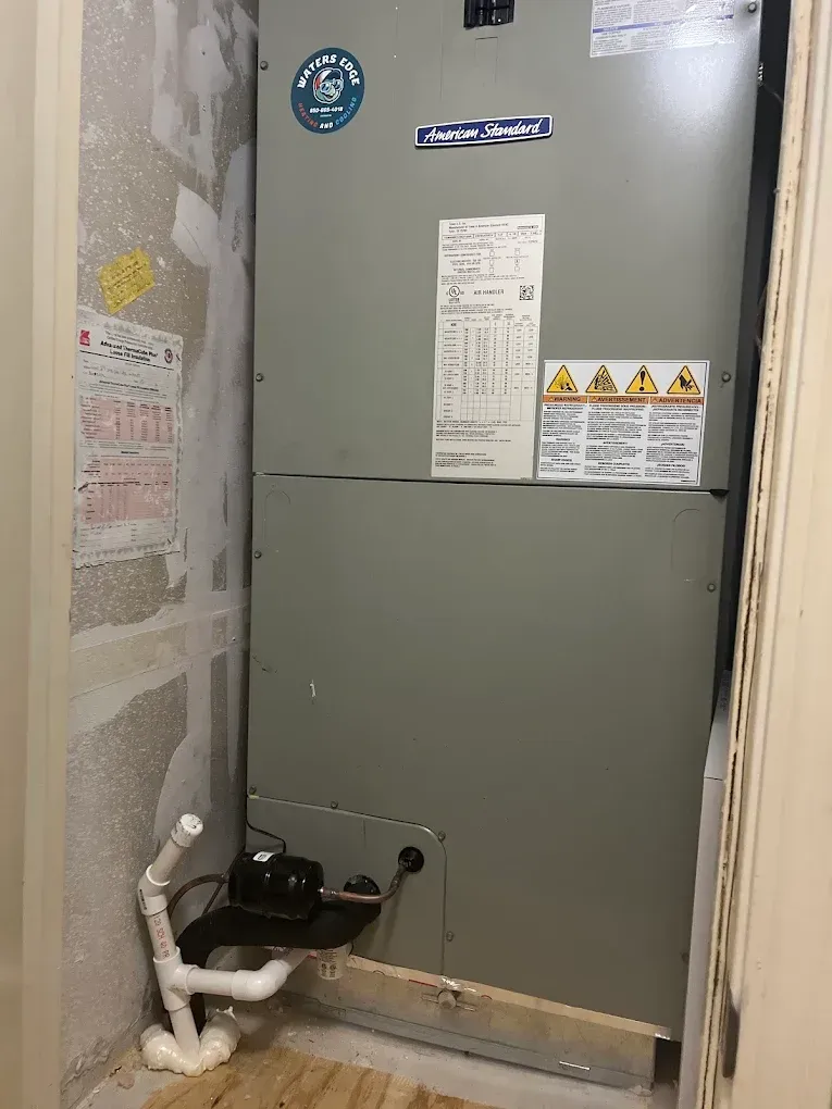 Gray air handler unit in a closet with exposed drywall and drainage pipes.