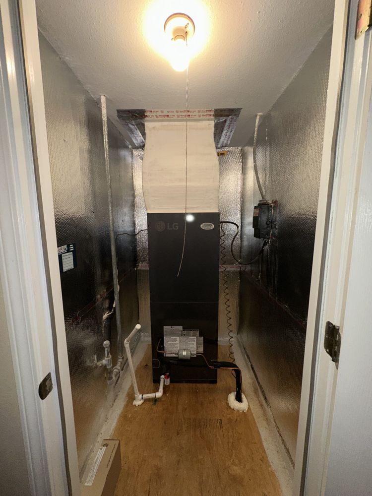 A furnace in an enclosed closet with silver insulated walls, light fixture, and door frames.