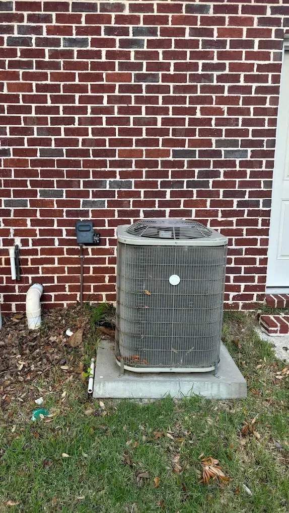 Air conditioning unit outside brick building. Gray unit on concrete pad, green grass, electrical box.
