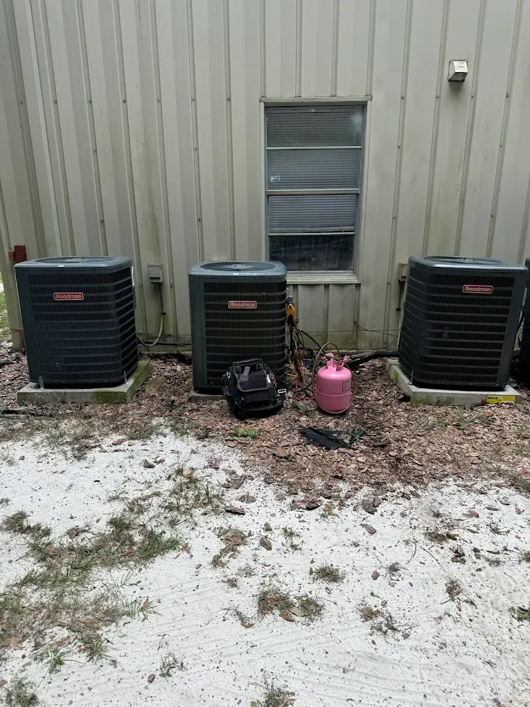 Three black HVAC units outside a building with a window; a black bag and pink tank are nearby.