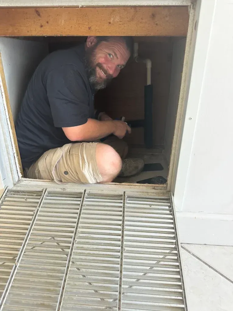 Man works on plumbing, smiling, inside a cabinet with a metal grate.