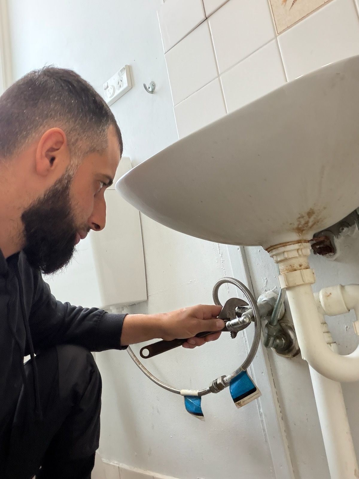 Person repairing a white sink faucet in a tiled bathroom with a wrench — Start 2 Finish Plumbing in Yagoona, NSW