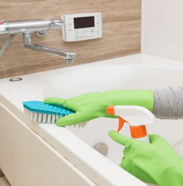 A person wearing green gloves is cleaning a bathtub with a brush and spray bottle.