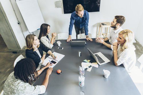 Group of people in a meeting around a table, one standing and speaking.