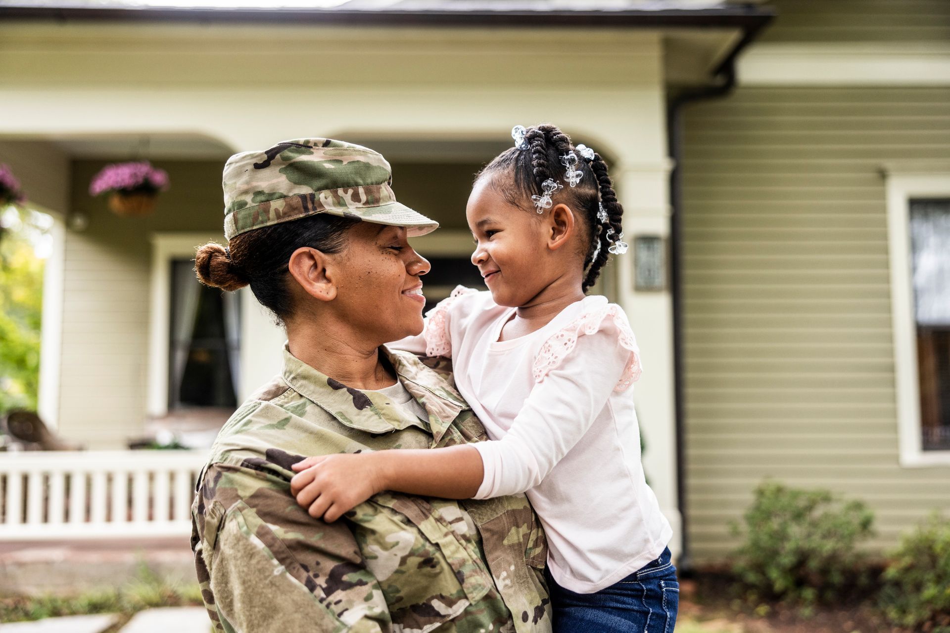 Soldier in uniform holds a smiling child outside a house.