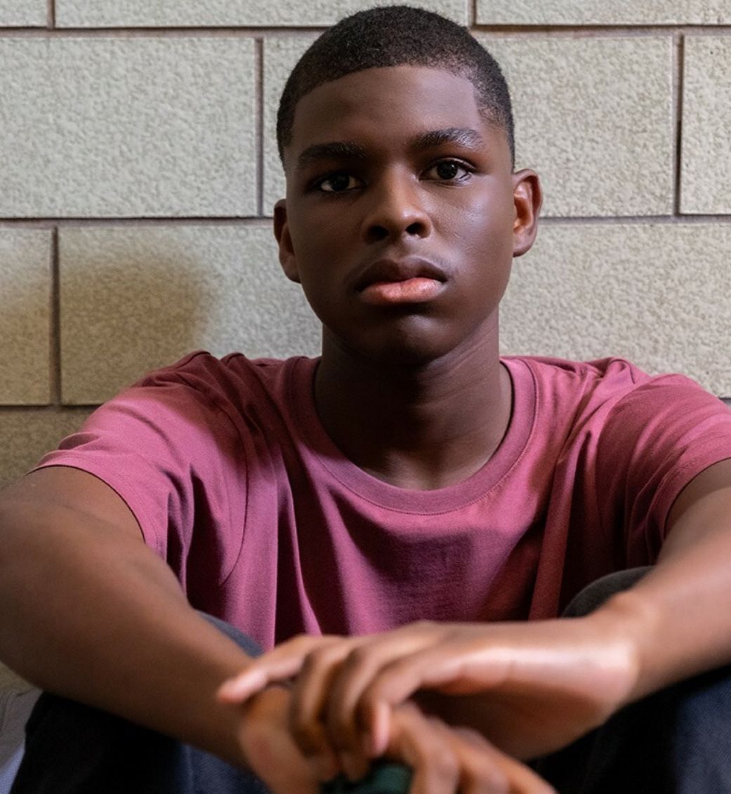 Teenage boy, in a white shirt, looks down with concerned expression; another person's hand on his shoulder.