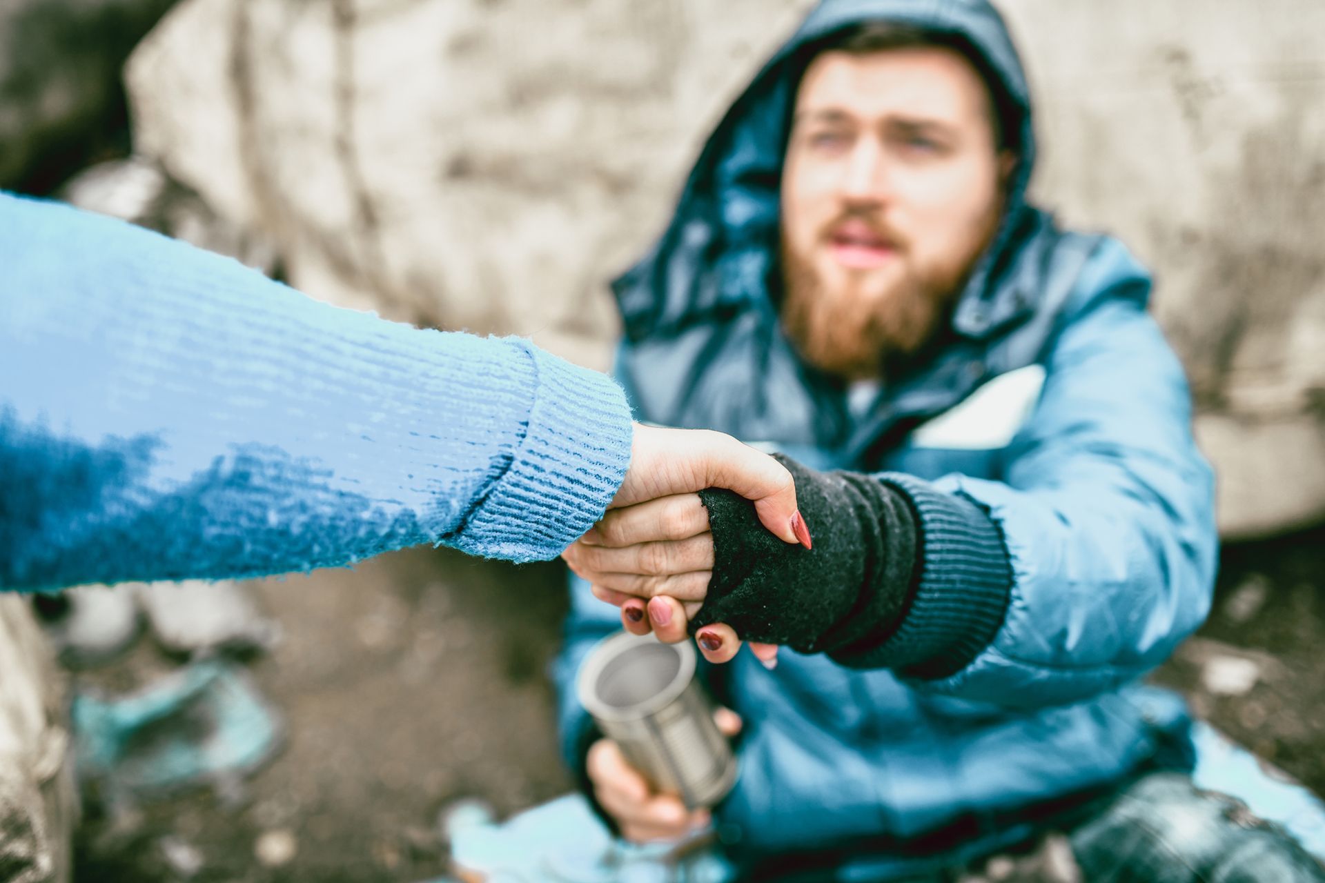 Person in blue jacket shakes hands with someone wearing blue sweater, holding a metal cup outside.