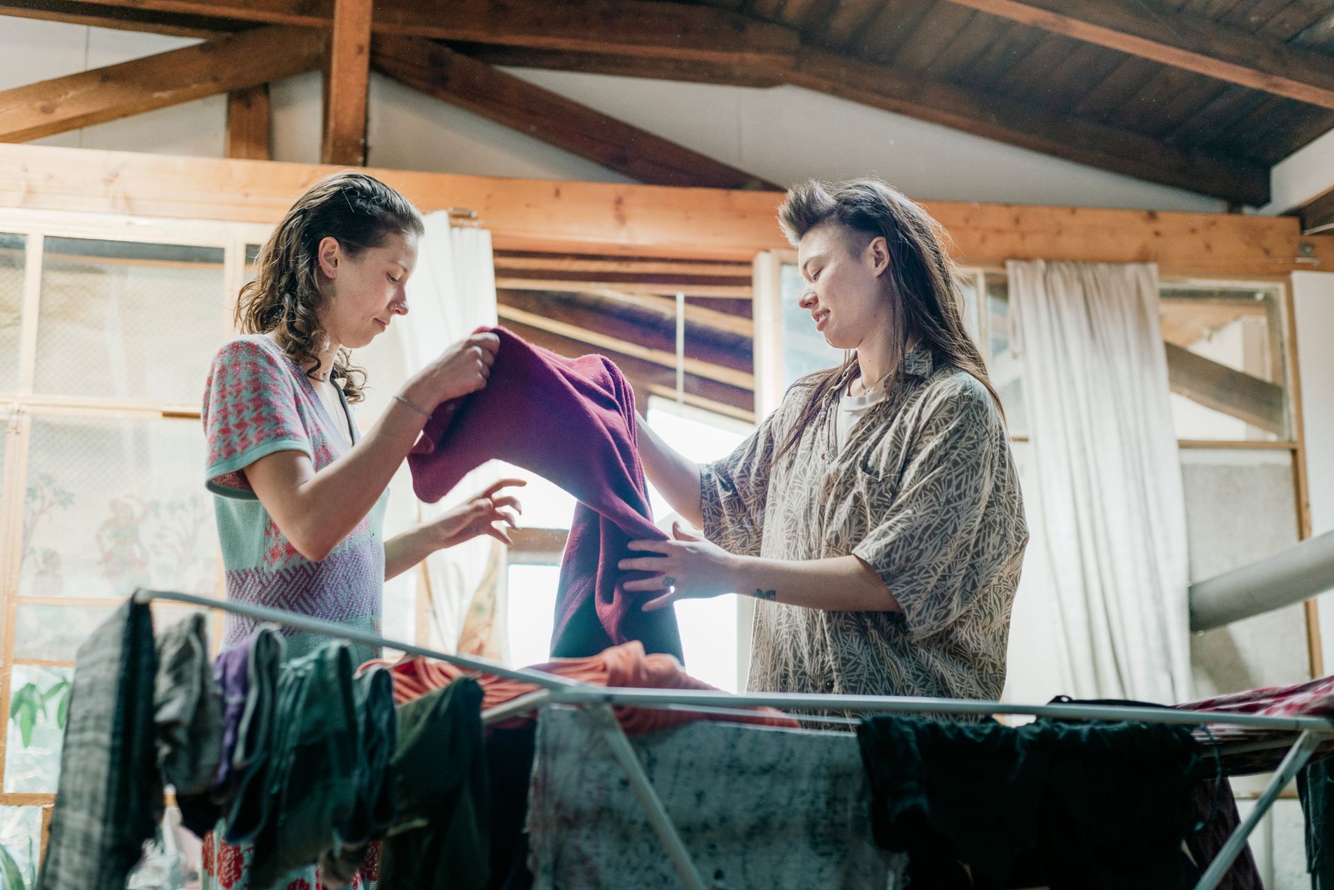 Two women folding laundry indoors; one with curly hair, the other with dreadlocks.