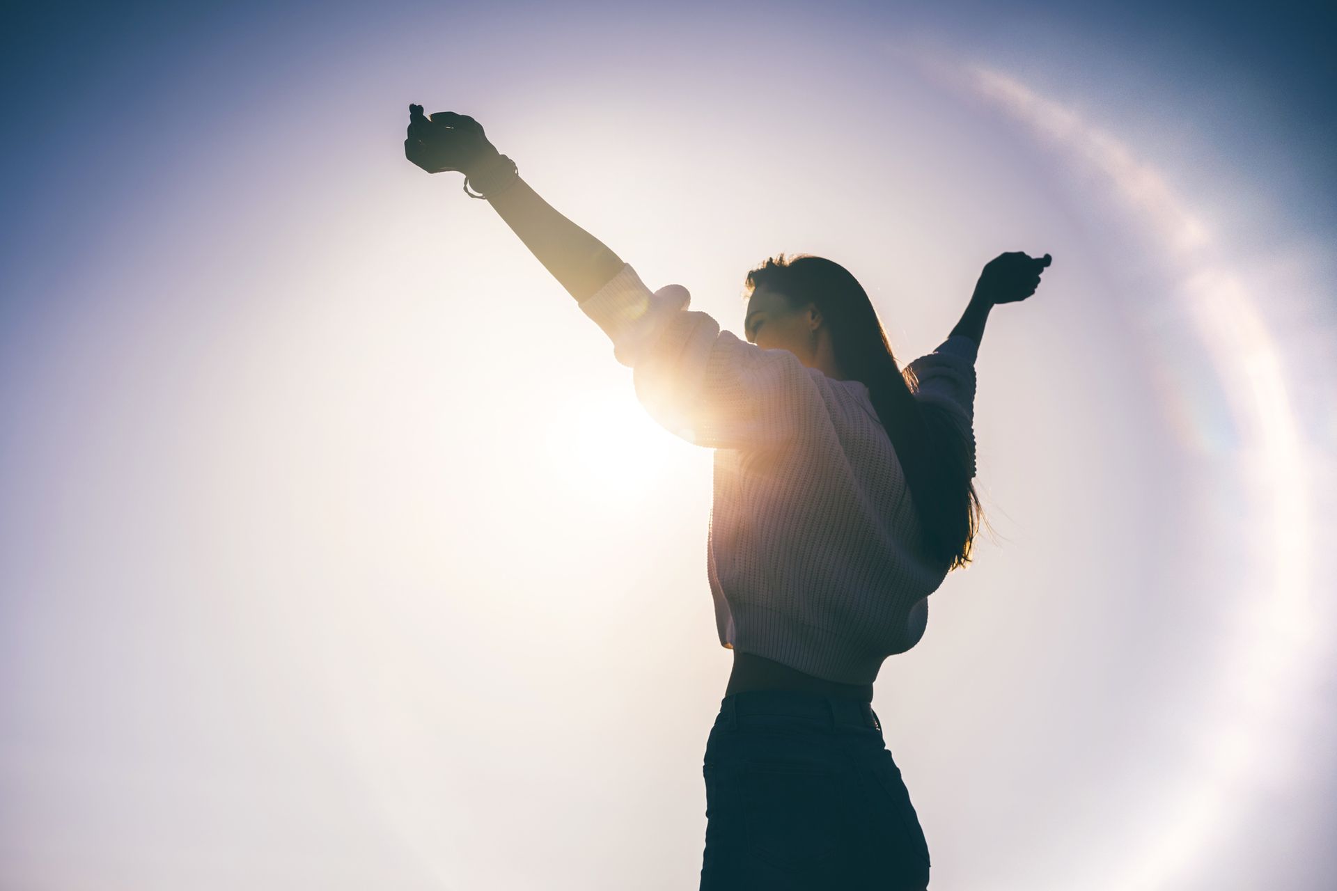 Woman with arms raised, silhouetted against bright sun with a halo effect.