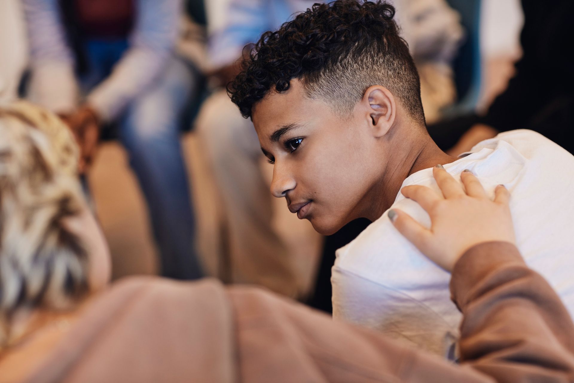 Teenage boy, in a white shirt, looks down with concerned expression; another person's hand on his shoulder.