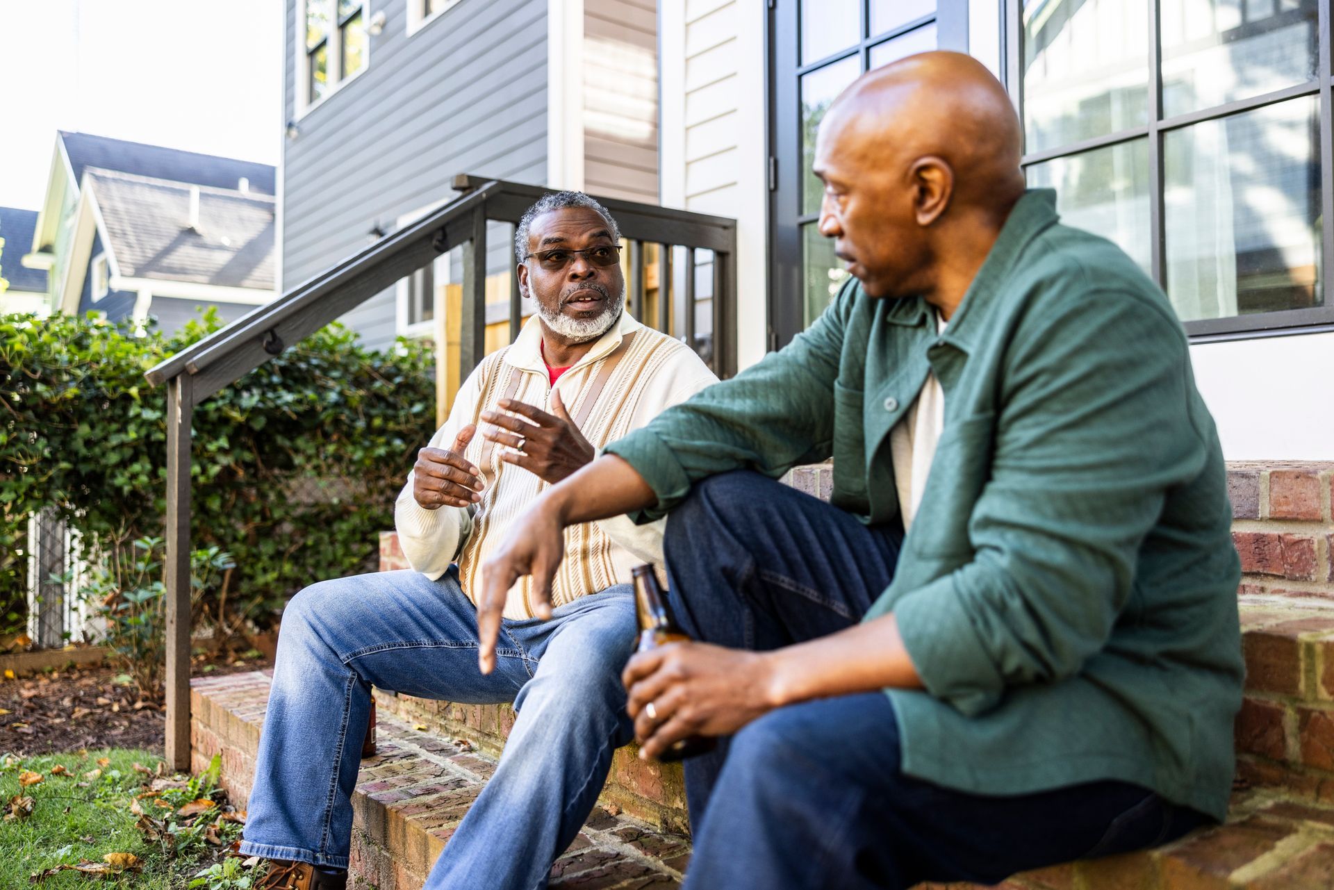Two Black men talking and sitting on steps outside a house. One holds a beer, gesturing.
