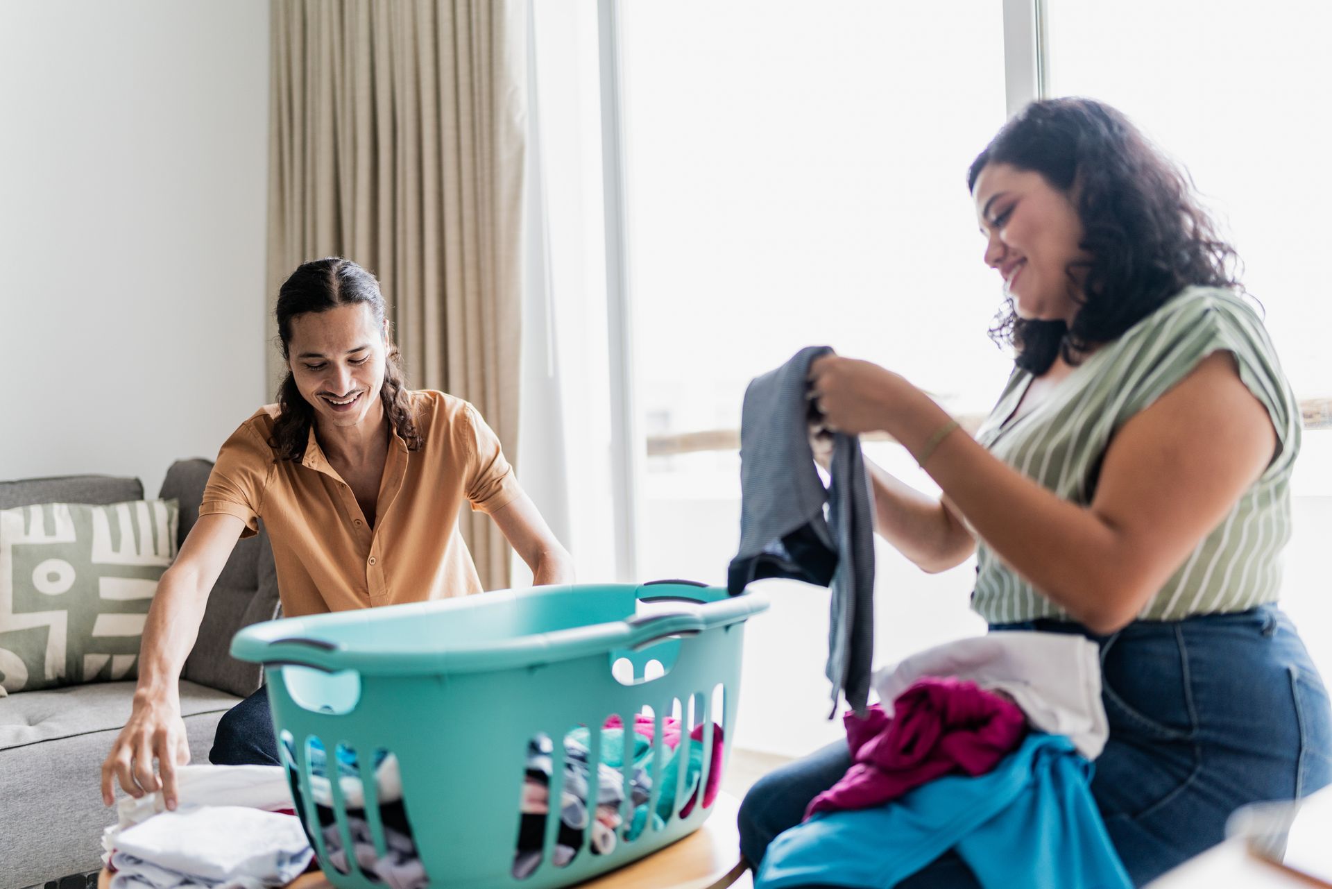 Two women folding laundry together in a light-filled room, smiling. One is holding a dark shirt.