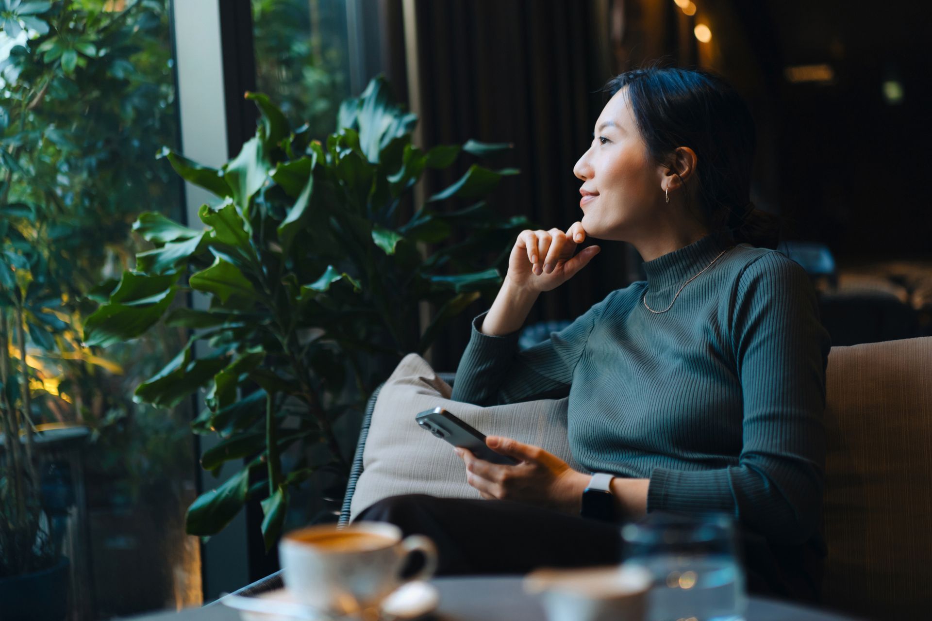 Woman gazing out a window, holding phone, in a dimly lit room with a plant and coffee cup.