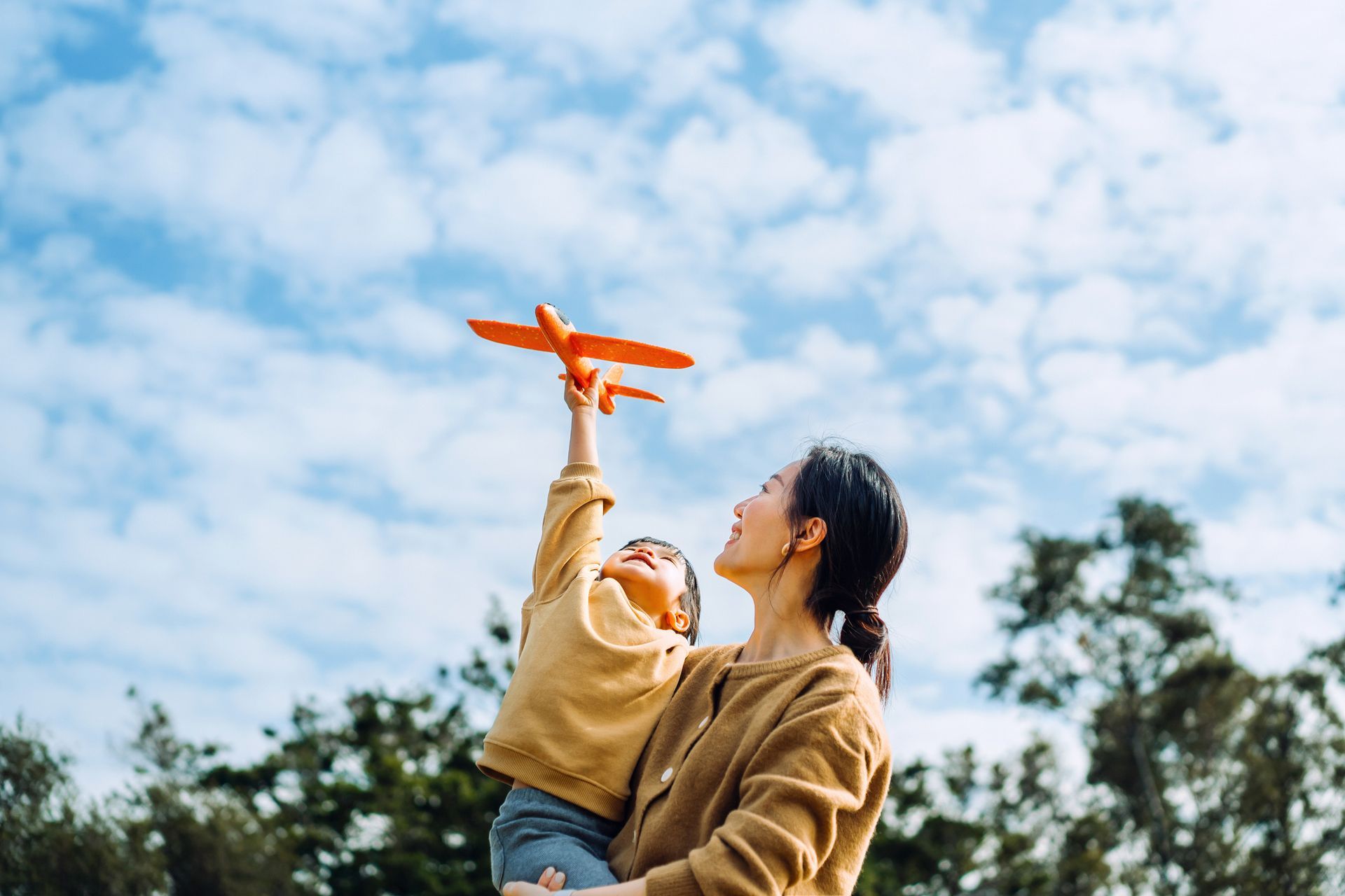 Woman holding a child, raising a toy airplane against a blue sky with clouds.