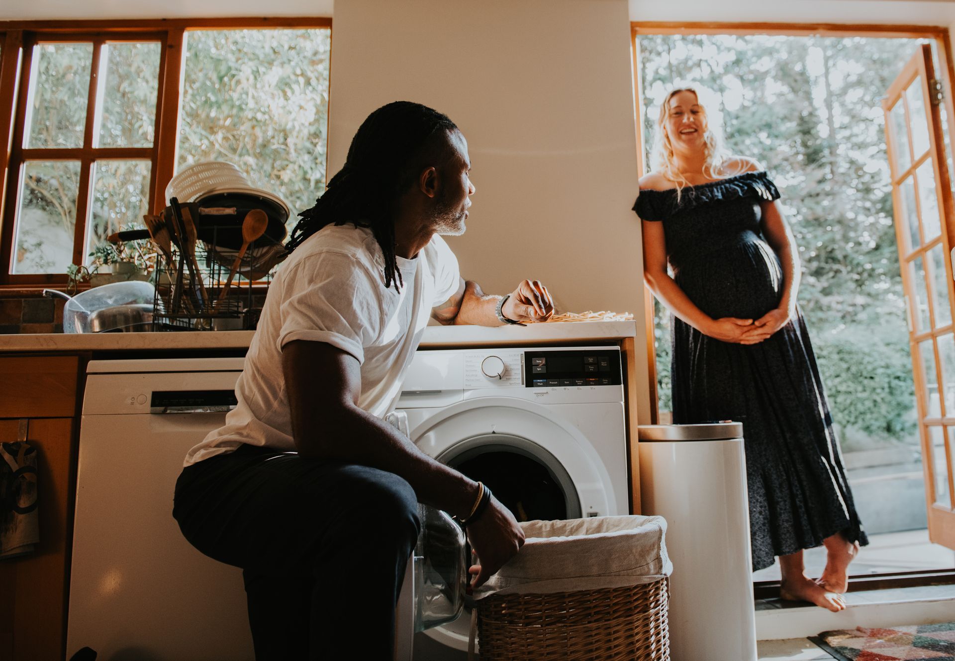 A pregnant woman and a man in a laundry room, looking at each other, one holding a basket.