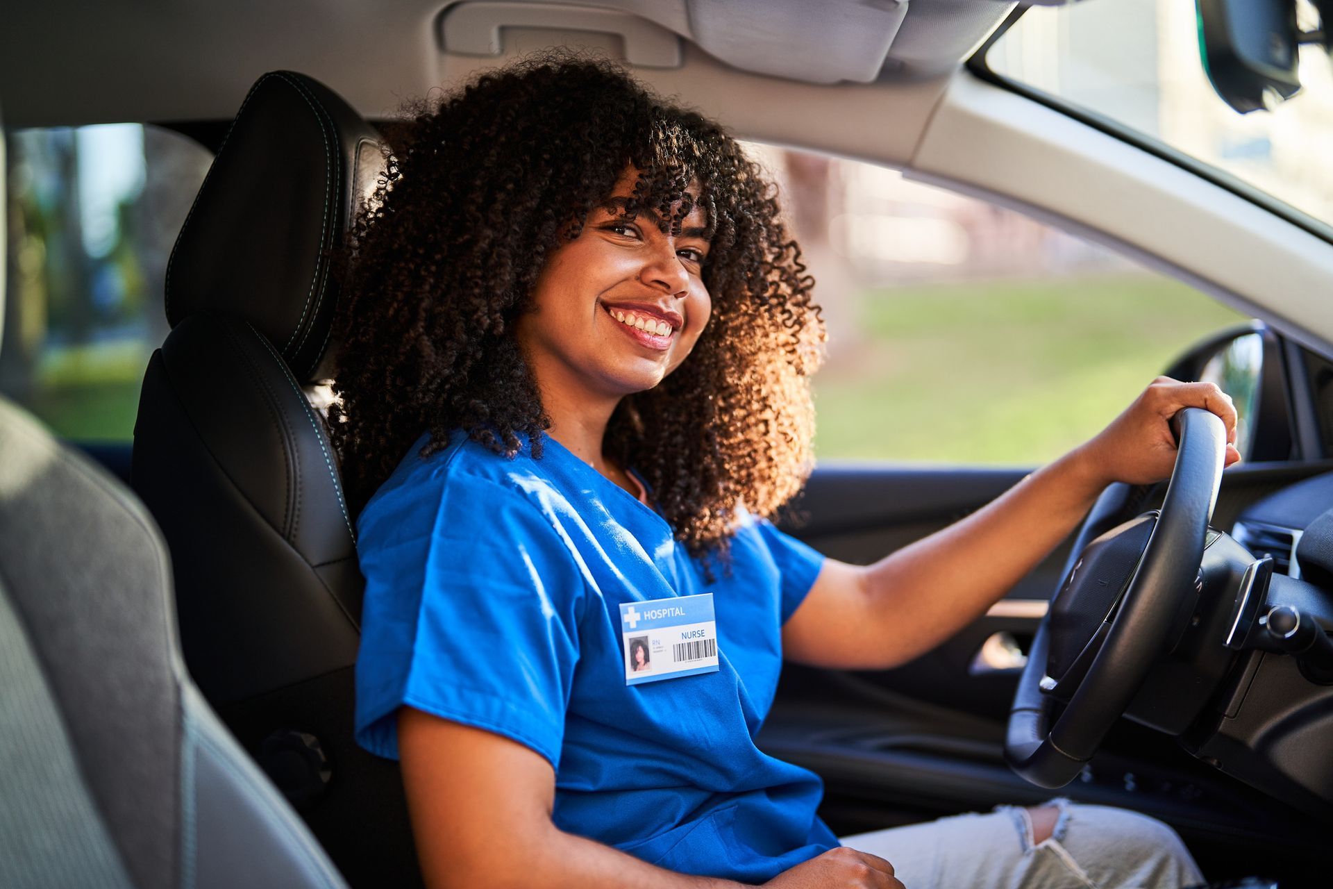 Smiling woman with curly hair, in blue scrubs, driving a car.