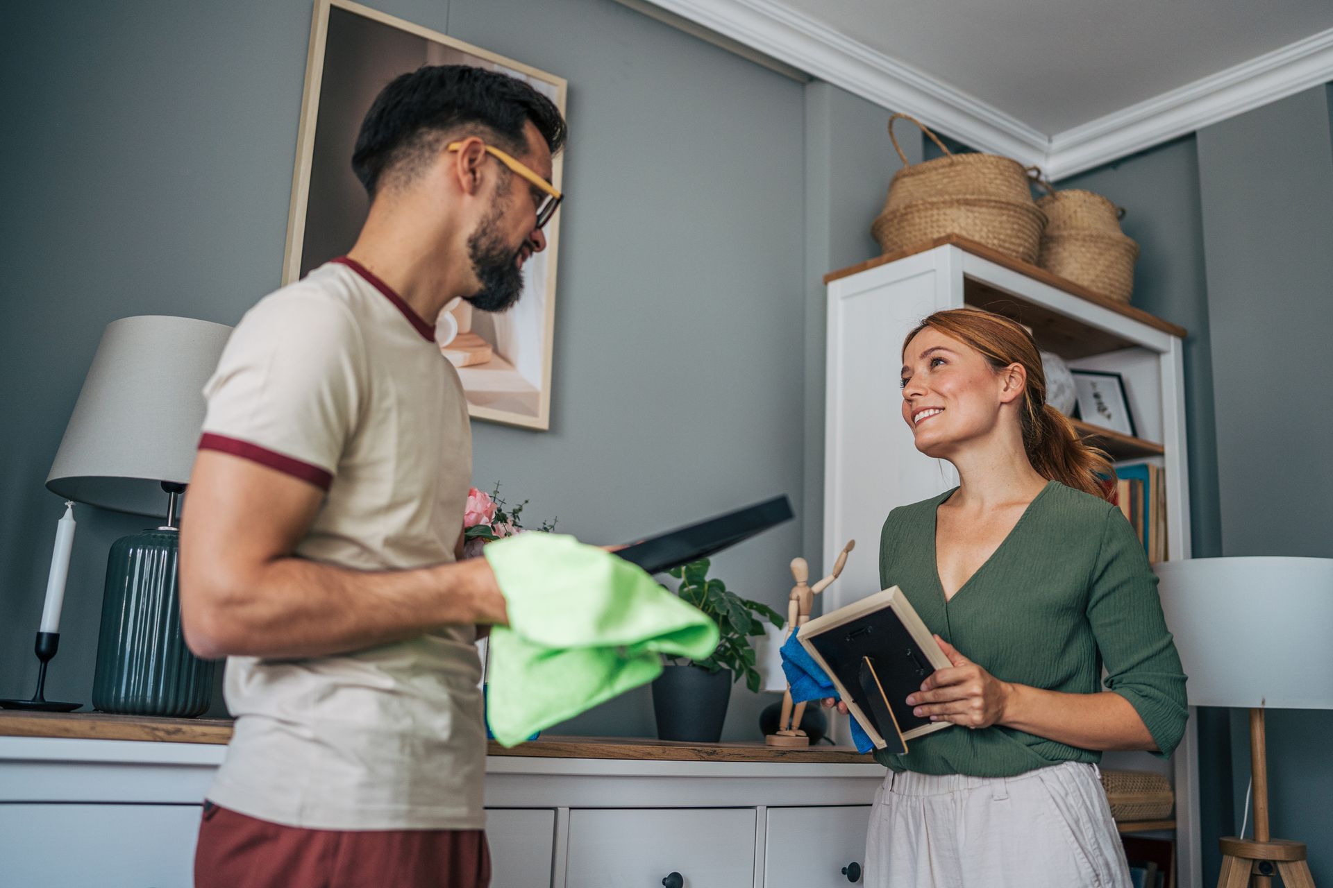 Man and woman cleaning a room, smiling. Man holds a green cloth. Woman holds a framed picture.