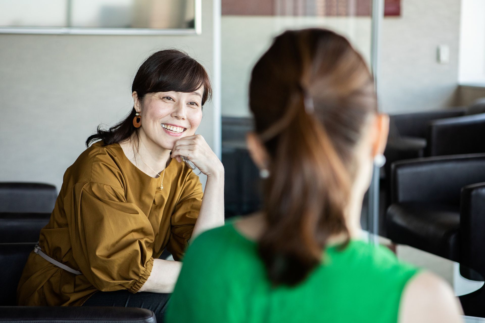 Woman in brown top smiles at another woman in a green top, in a modern setting with dark chairs.