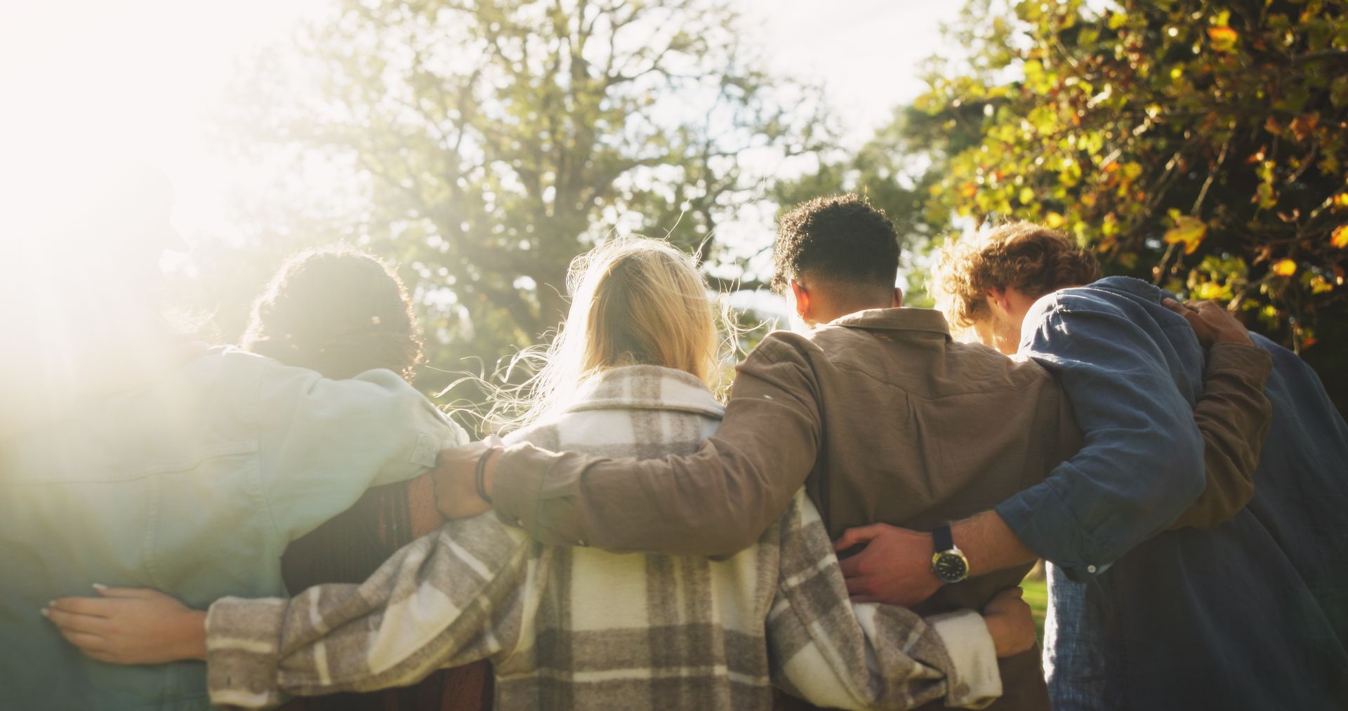 Group of five people with arms around each other in a park, backlit by the sun.