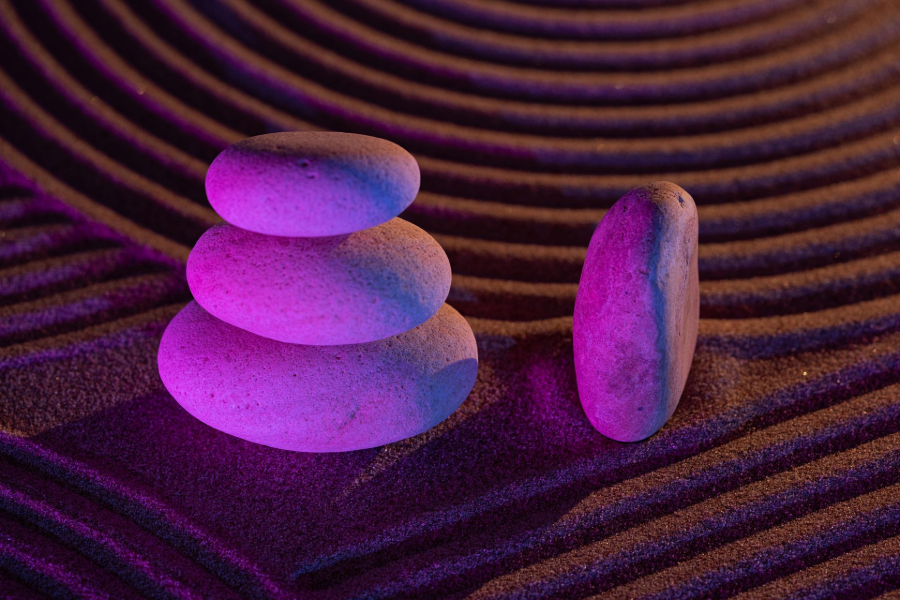Three stacked smooth stones and a standing stone on rippled sand, lit in purple and pink.