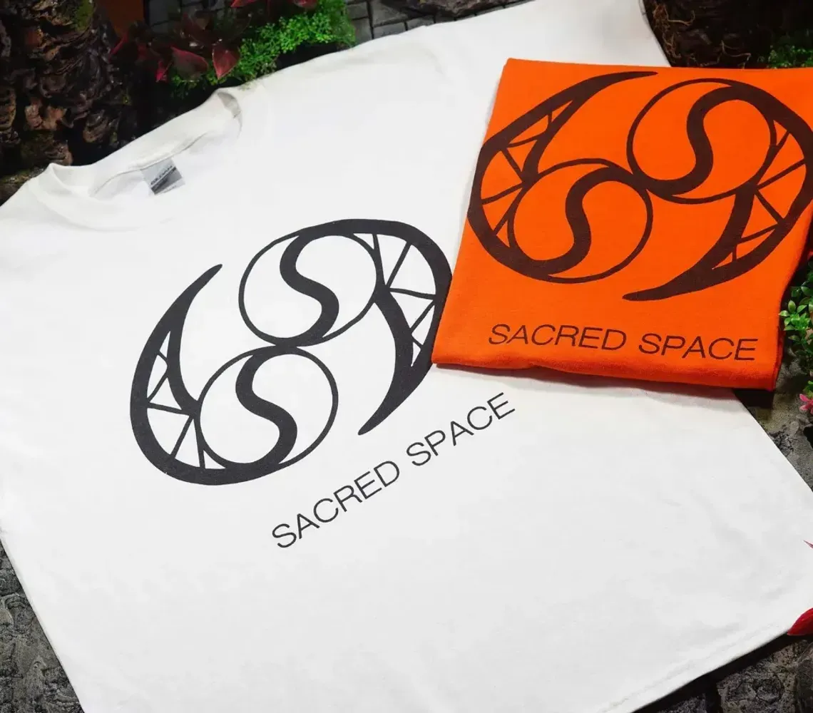 White T-shirt and orange card featuring a black circular logo with “SACRED SPACE” text on foliage background
