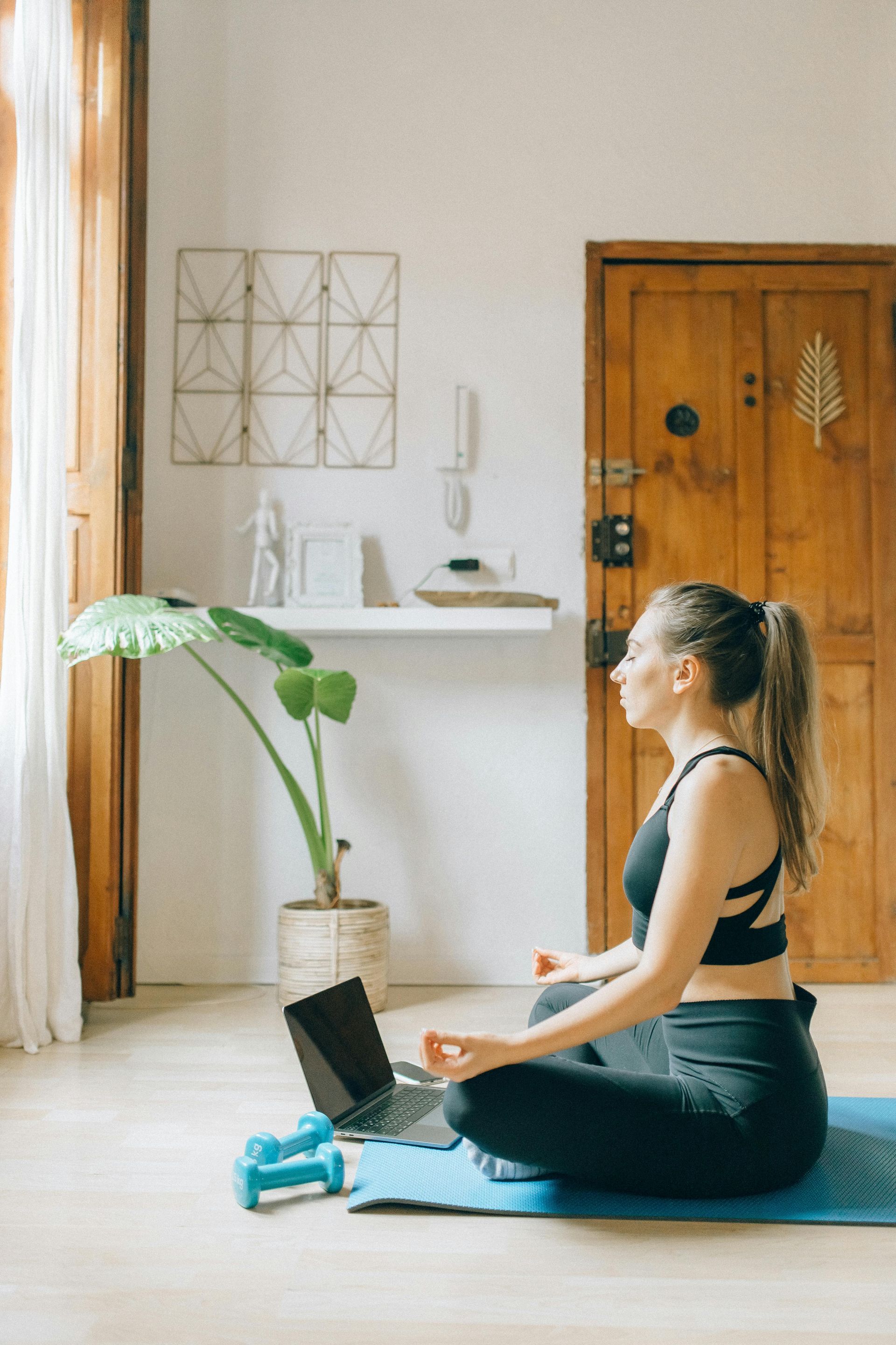 Woman practicing seated meditation on a yoga mat beside a laptop in a bright room