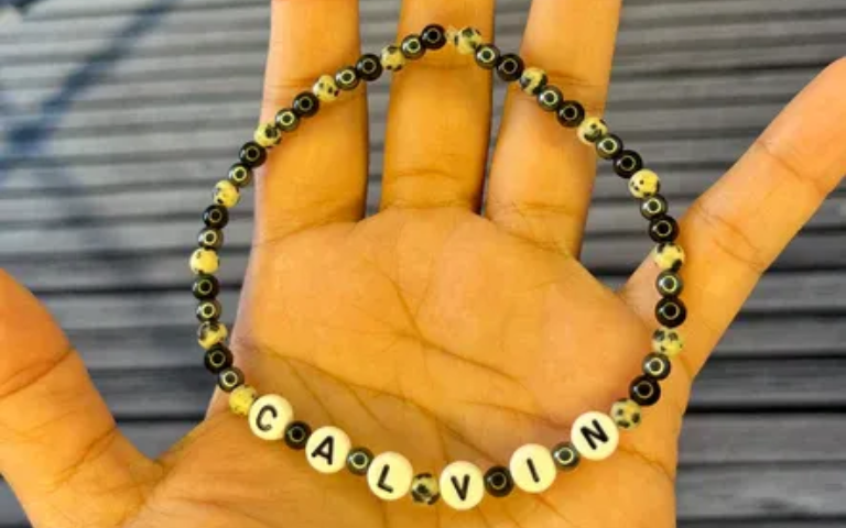 Hand holding a beaded bracelet spelling “CALVIN” on a gray wooden surface