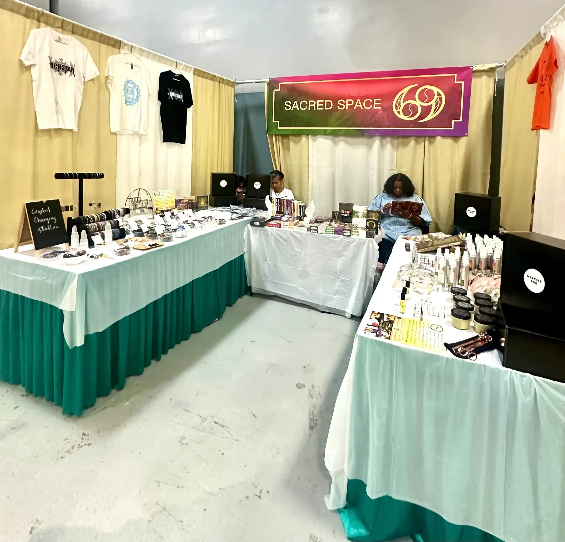 Indoor vendor booth with hanging T-shirts, tables of crafts, and a “Sacred Space” banner in the back