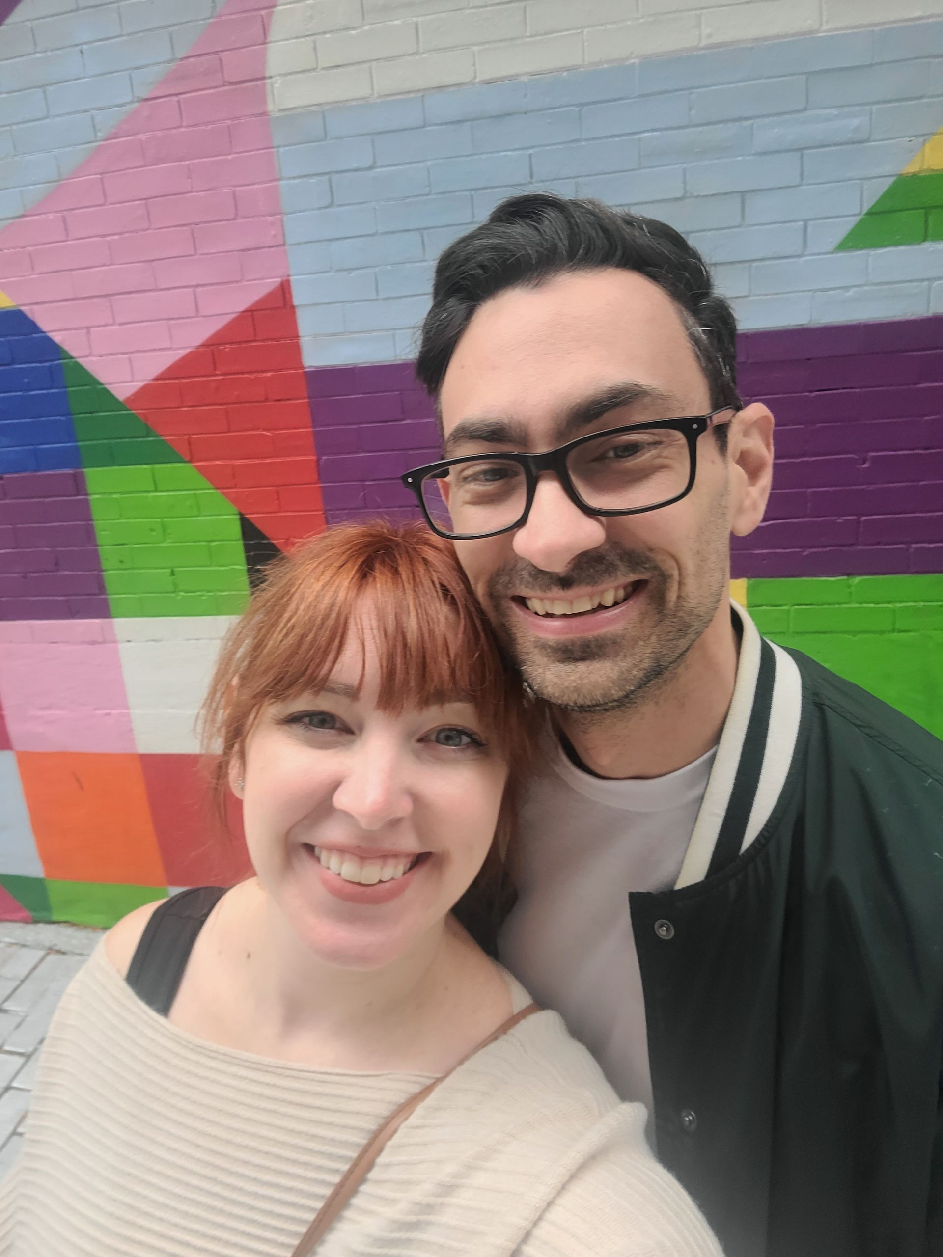 Couple smiles for a selfie at a lake with mountains in the background.