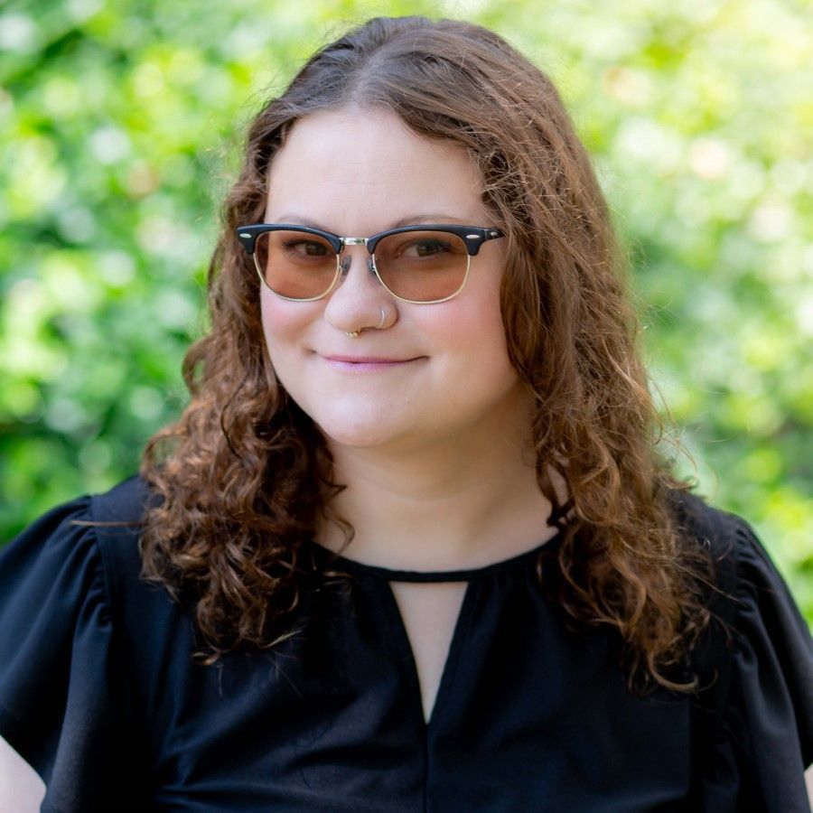 Woman with curly brown hair and glasses smiles, wearing a peach-colored button-down shirt.