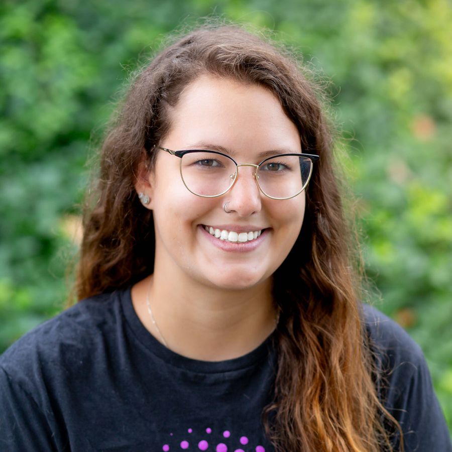 Woman with glasses smiles, outdoors, wearing black shirt with purple graphic, long brown hair.