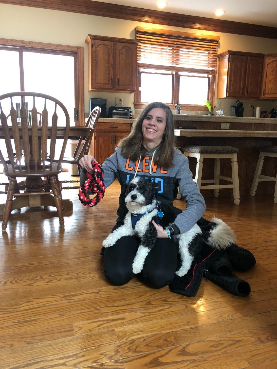 Woman kneeling with a black and white dog, holding a leash. Kitchen setting, wood floor and cabinets.