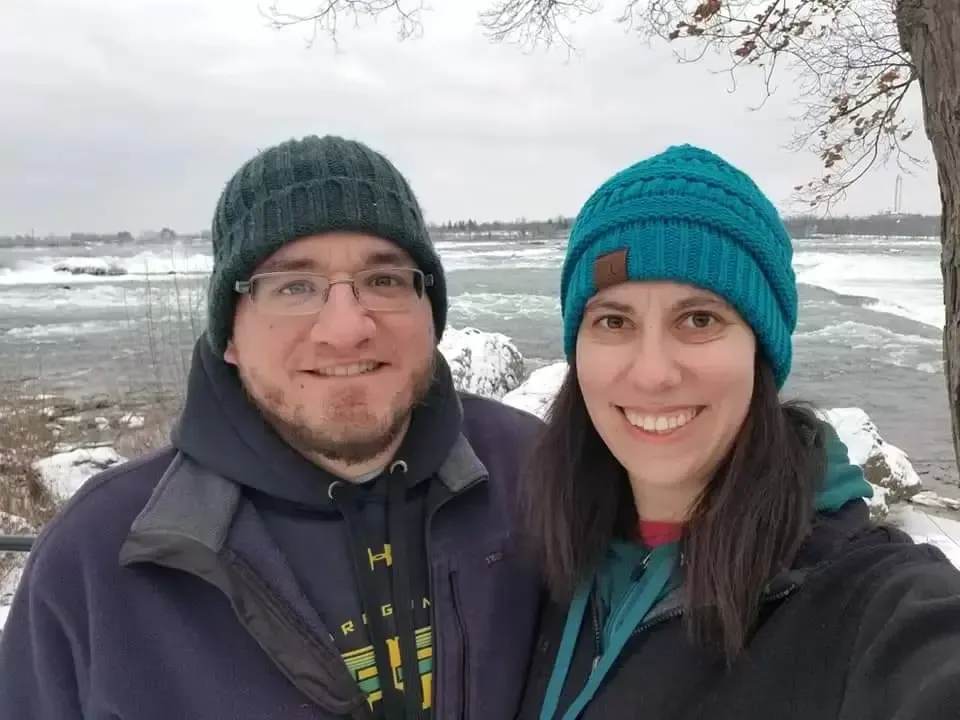 Couple in winter hats smile in front of a snowy river.