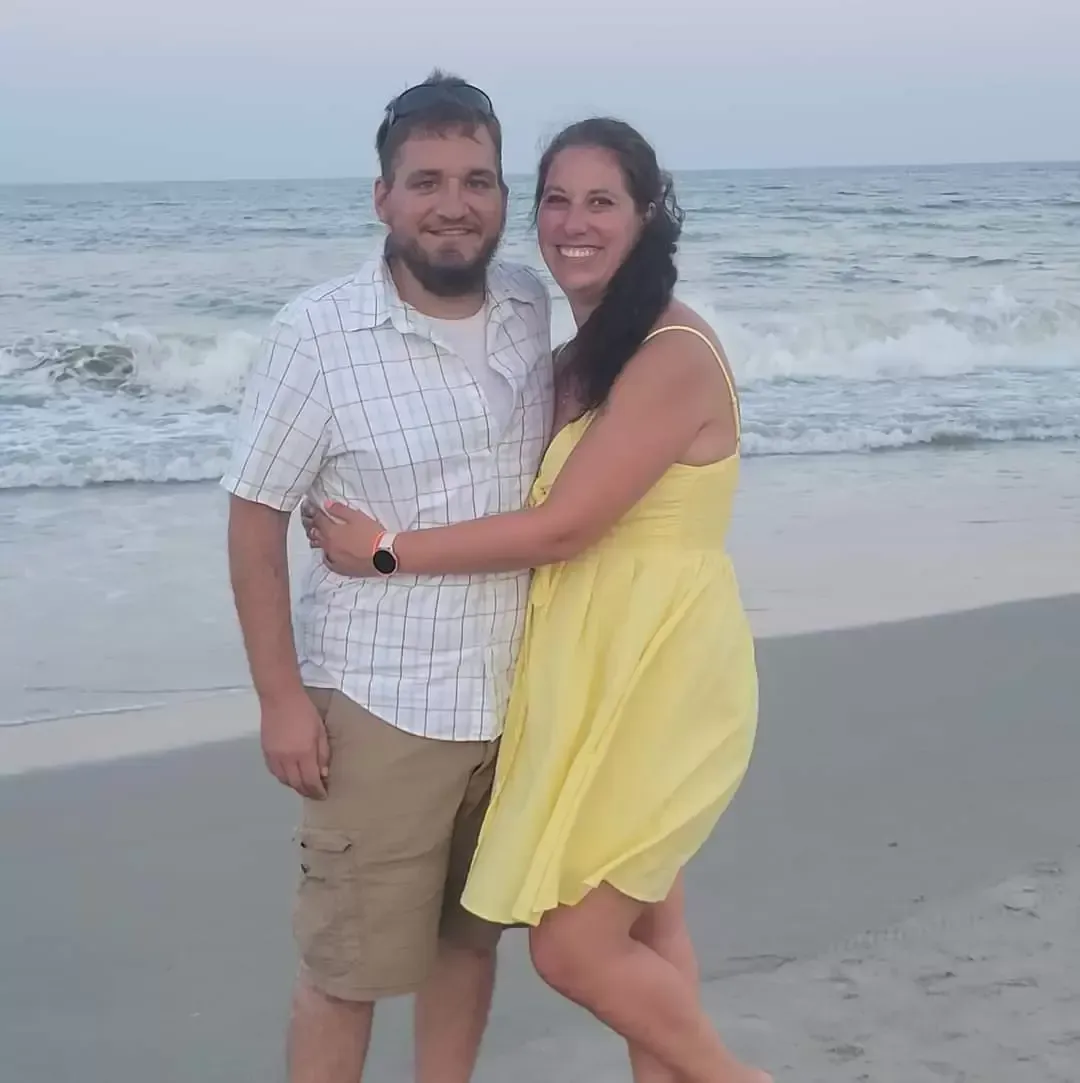 Couple embracing on a beach; man in tan shorts and plaid shirt, woman in a yellow dress. Ocean in background.