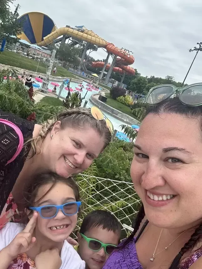 Four people smiling at a water park. Two women and two children in front of a lazy river and water slides.
