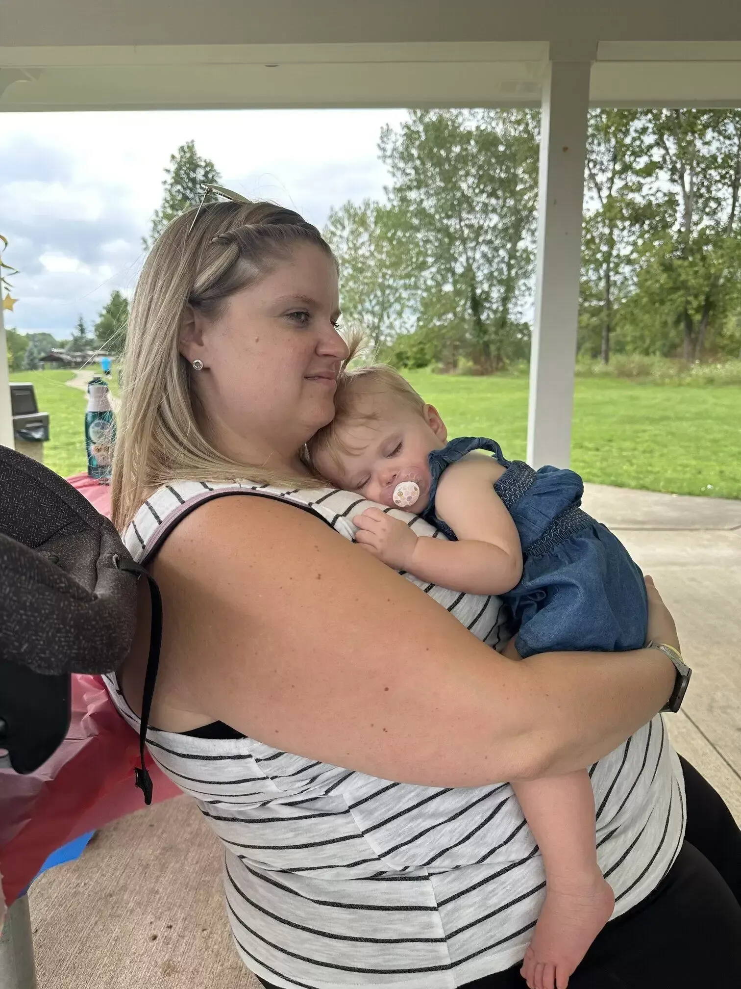 Woman in striped shirt holds sleeping baby in denim outfit outside.