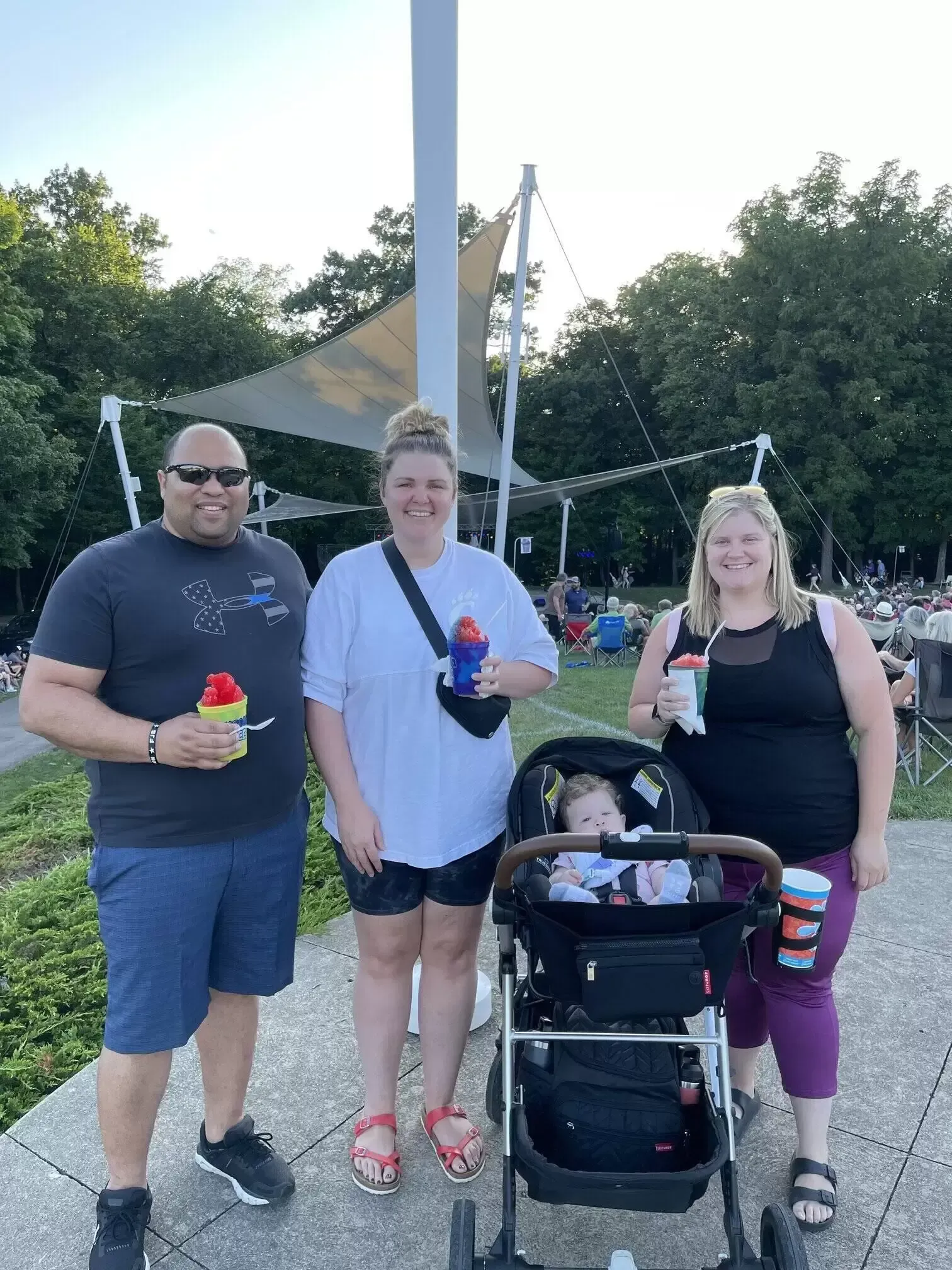 Three adults and a baby in a stroller at an outdoor event, holding snacks, with park and shelter in the background.
