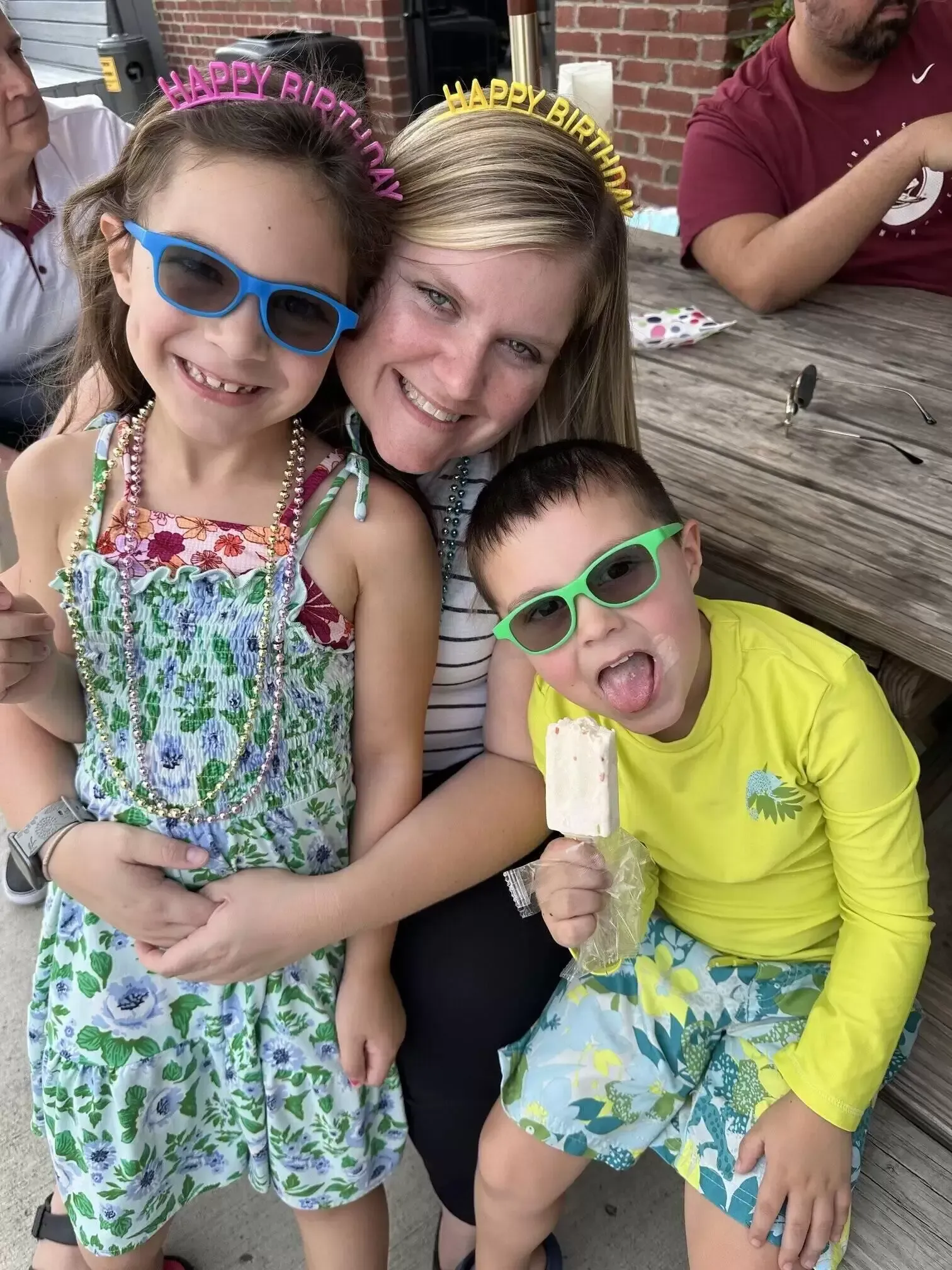 Woman embraces two children, all wearing sunglasses and birthday headbands outdoors.