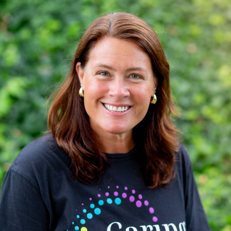 Woman with brown hair smiling, wearing a black shirt with a colorful logo. Green foliage background.